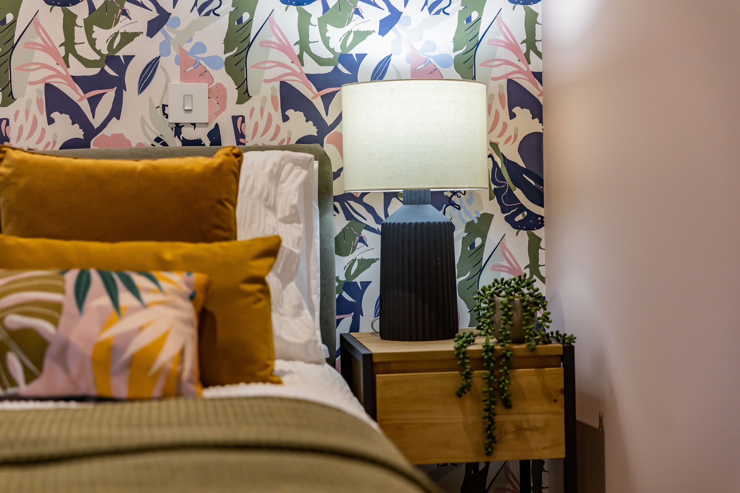 Bed with colourful pillows in the middle of a room next to a small wooden bedside table