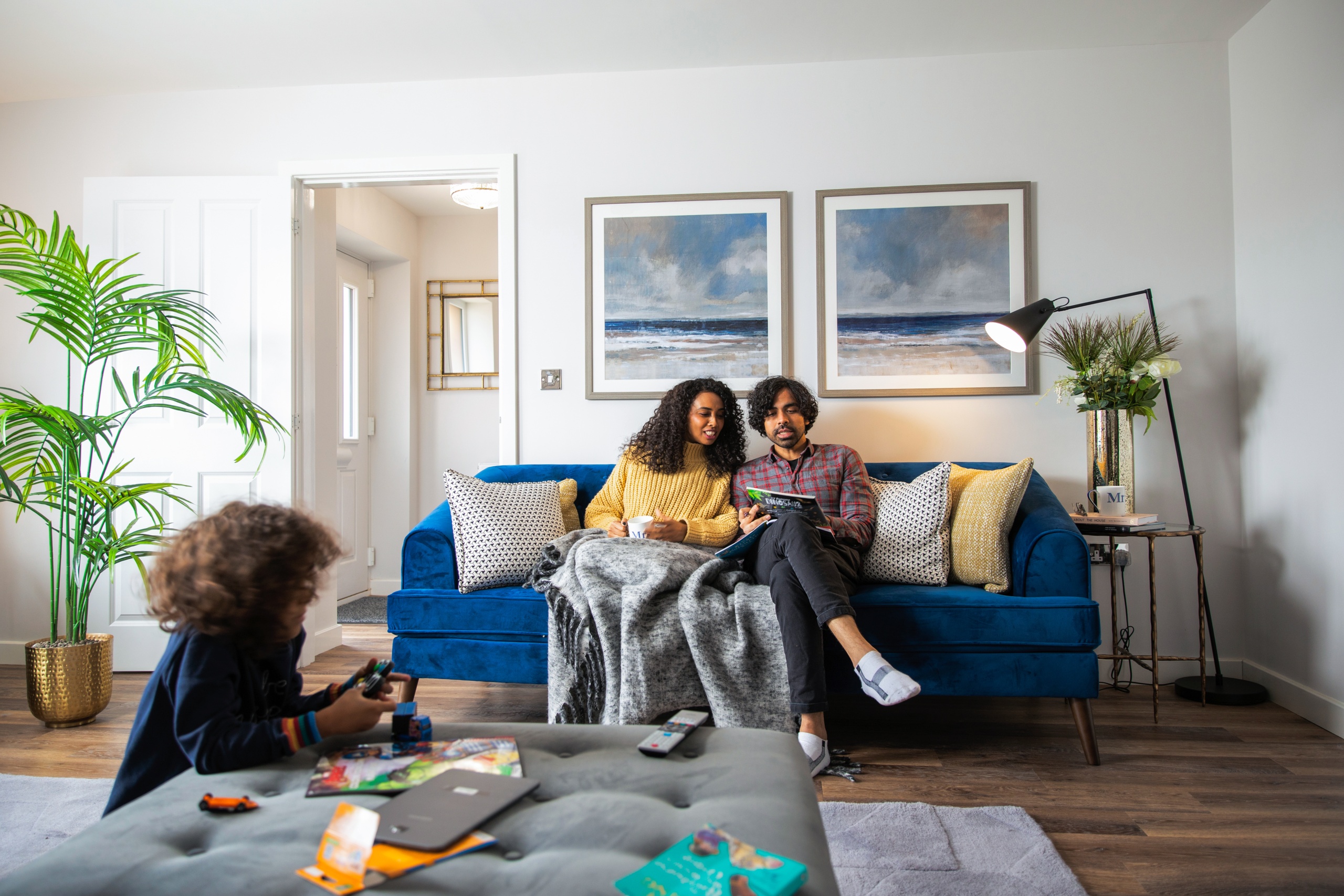 A young couple sat on the sofa looking at a magazine while their child plays with his toys in front of them