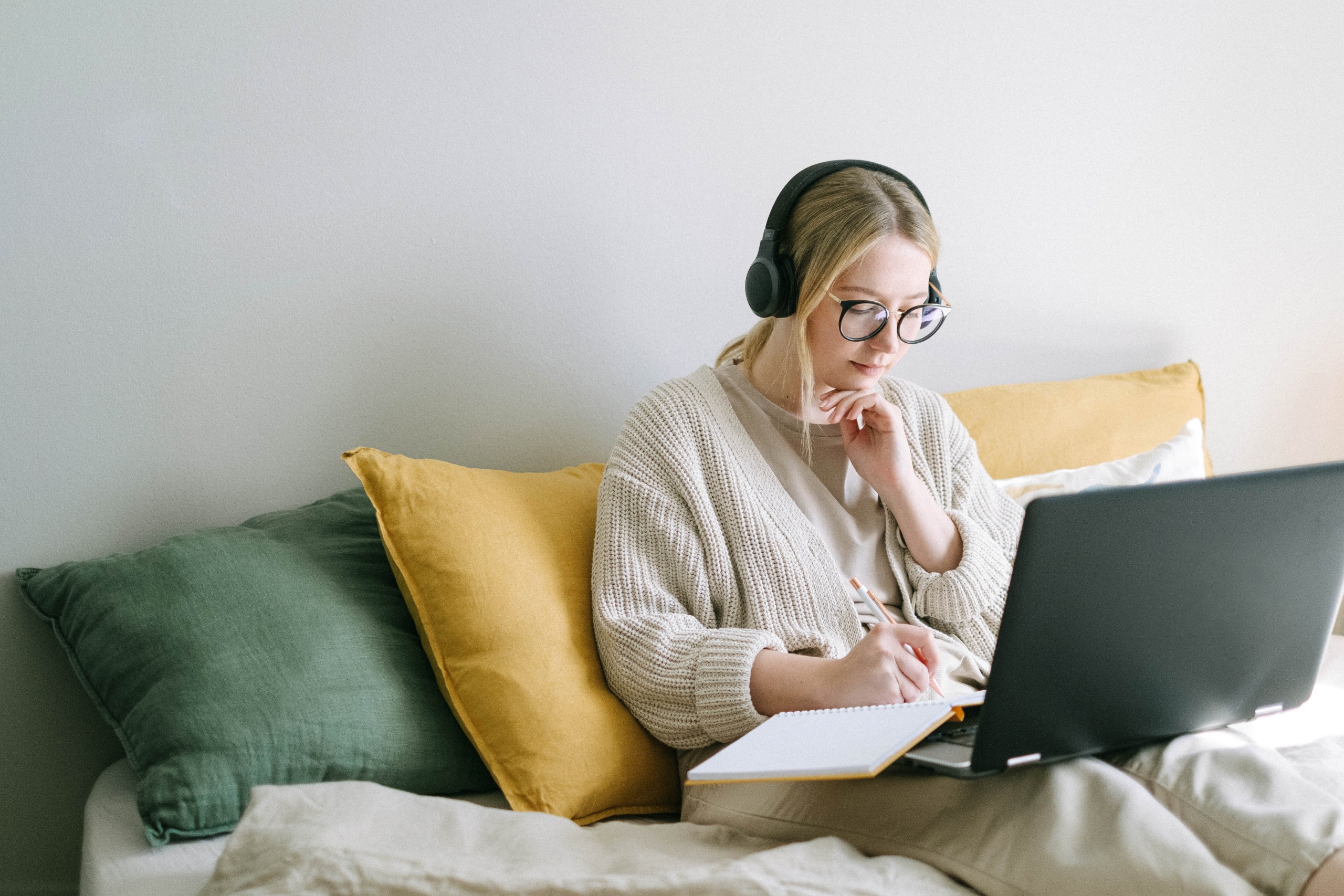 woman sat on her bed with headphones on taking notes off of her laptop